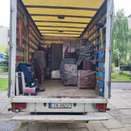 Wnętrze załadowanego busa transportowego z meblami zabezpieczonymi folią i kocami, widok z tyłu pojazdu na ulicę i budynek mieszkalny.