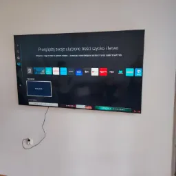Telewizor Samsung Smart TV zamontowany na białej ścianie, widoczne aplikacje streamingowe i kabel zasilający wpięty do gniazdka.