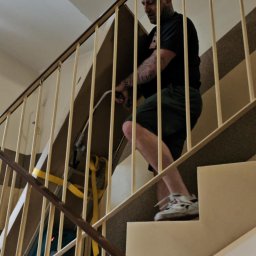 Przeprowadzki Transpot Piotr - Mężczyzna z tatuażami wnosi kartonowe pudło po schodach, używając wózka schodowego z żółtymi pasami. Widok z dołu, barierki schodowe w kadrze.