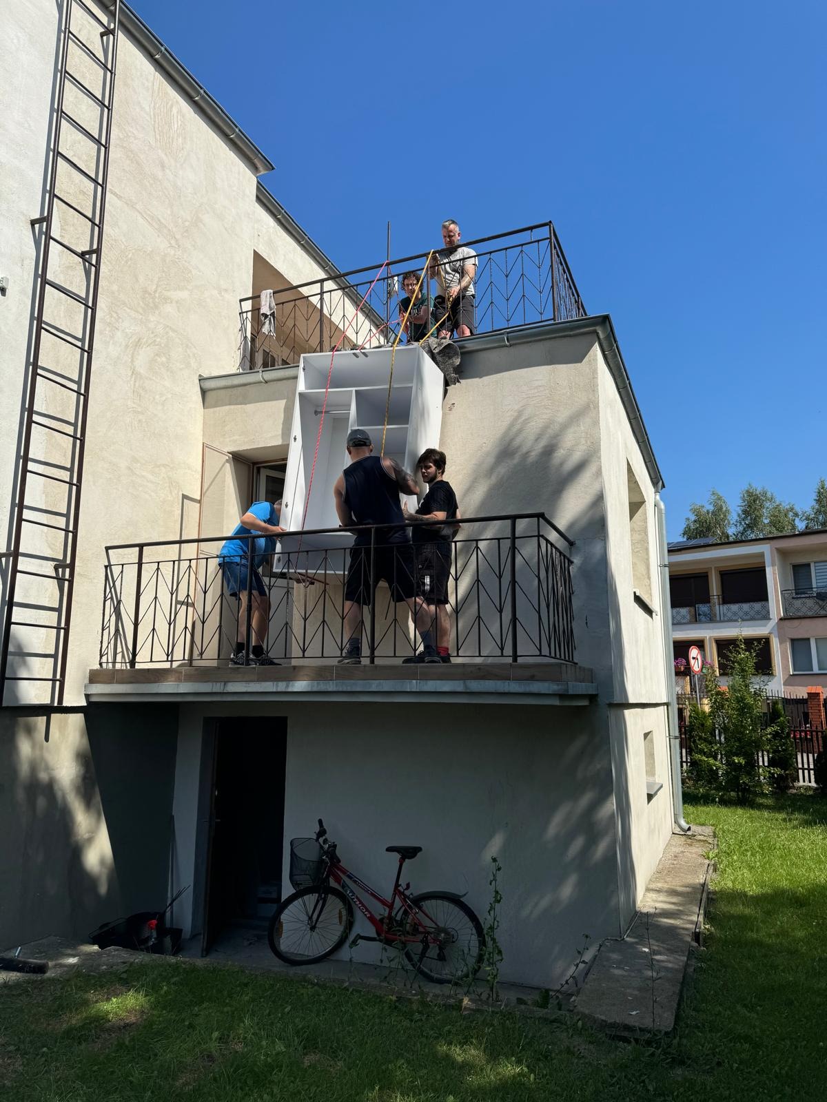 Czterech mężczyzn przenosi szafę z balkonu na balkon za pomocą lin, widok z dołu na dwupiętrowy budynek z rowerem opartym o ścianę.