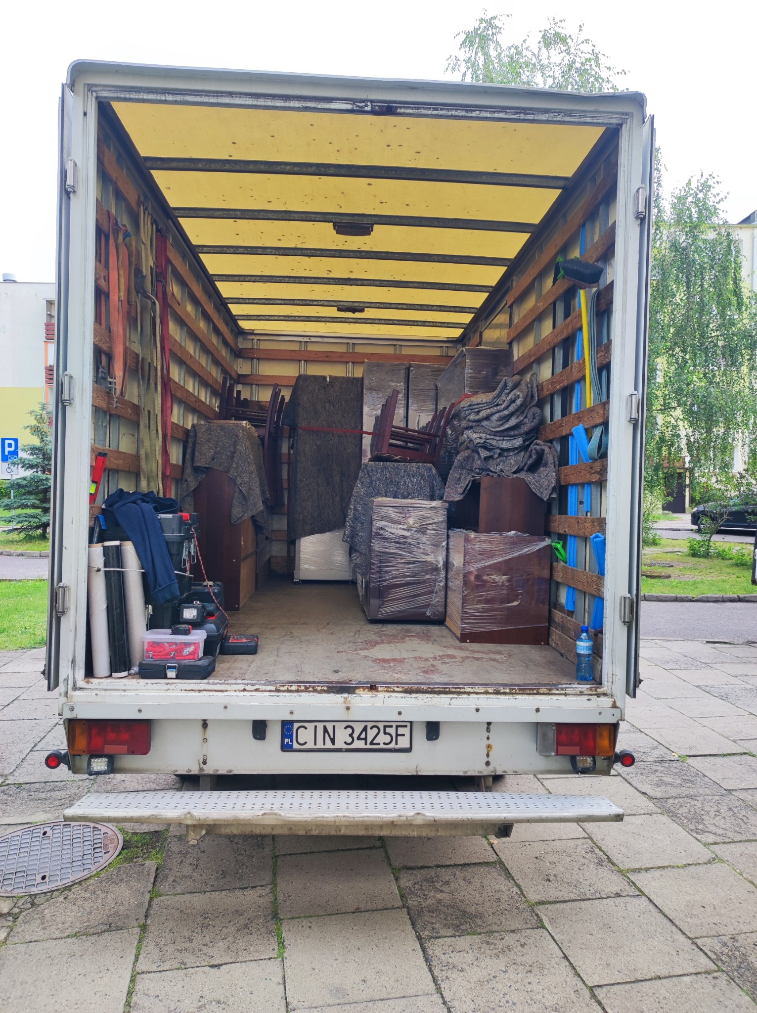 Wnętrze załadowanego busa transportowego z meblami zabezpieczonymi folią i kocami, widok z tyłu pojazdu na ulicę i budynek mieszkalny.