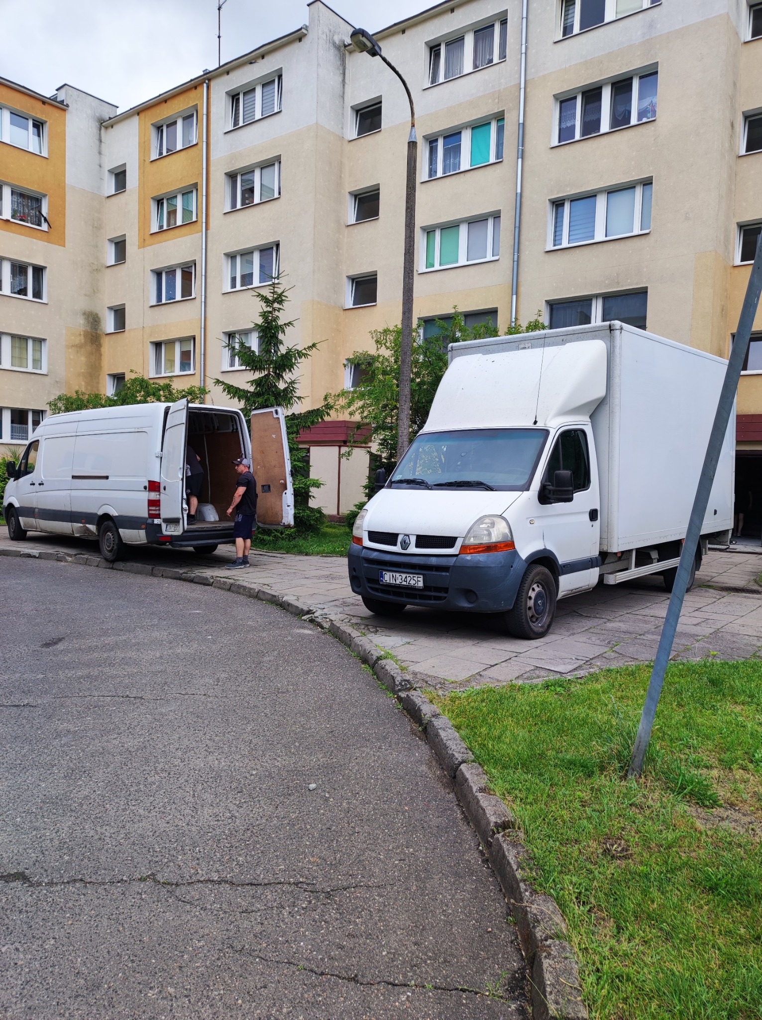 Dwa białe busy transportowe zaparkowane przed blokiem mieszkalnym podczas załadunku, jeden z otwartymi drzwiami i widoczną osobą w środku.