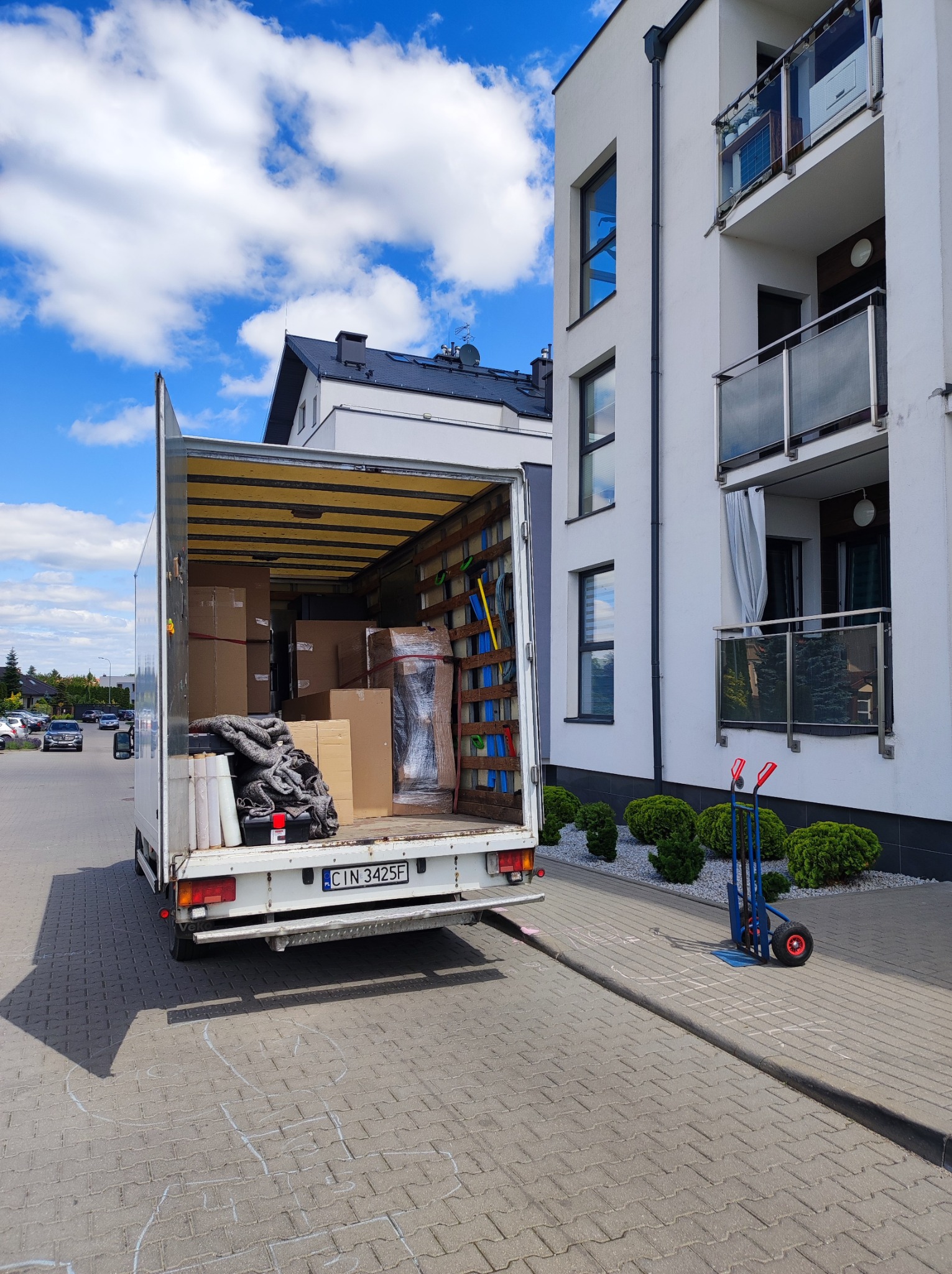 Otwarty tył białej furgonetki wypełnionej kartonami i zabezpieczeniami, zaparkowanej obok nowego budynku mieszkalnego z balkonami i wózkiem transportowym.