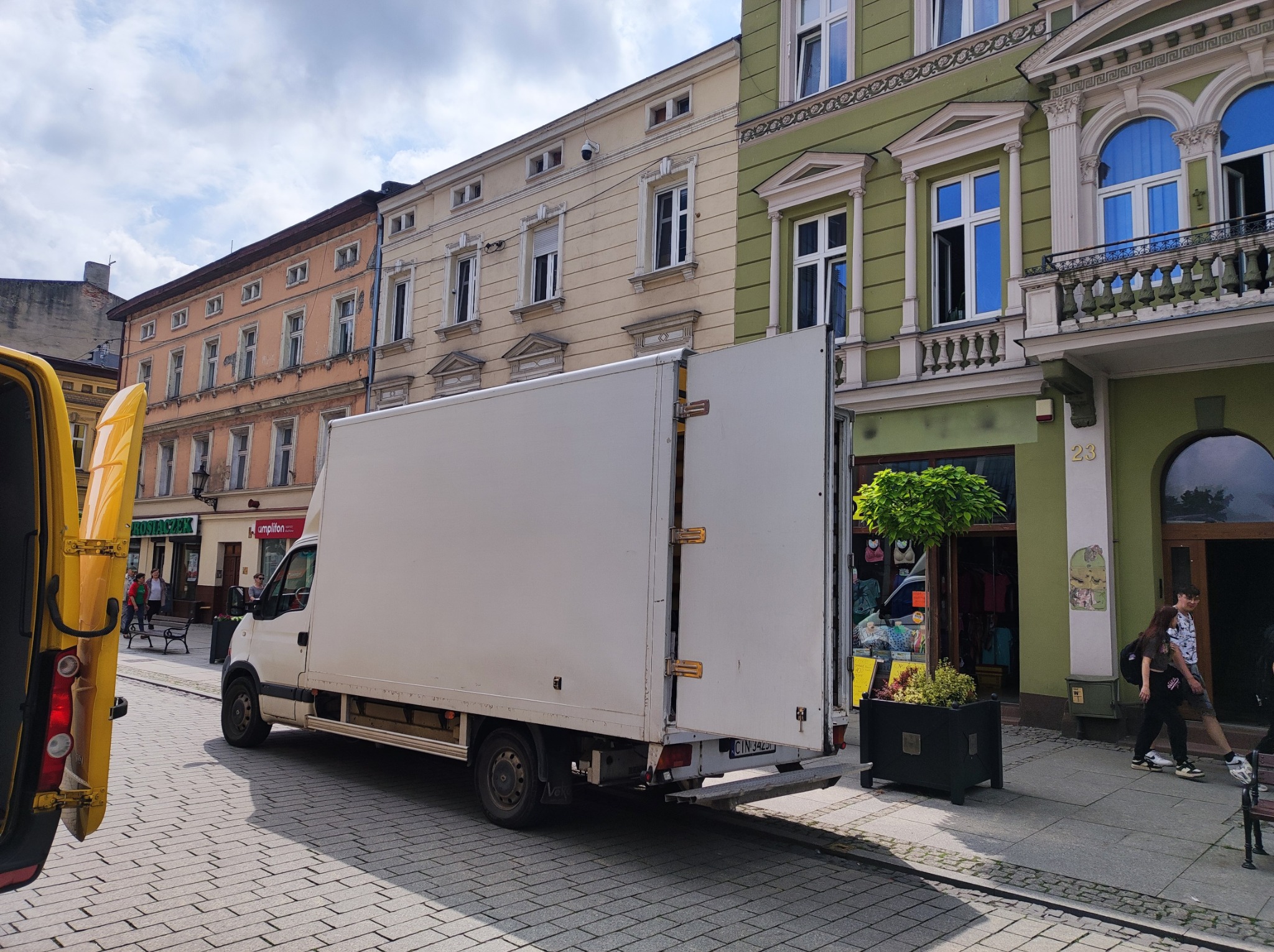 Biały samochód dostawczy z otwartymi drzwiami zaparkowany na ulicy w centrum miasta przed budynkami o zróżnicowanej architekturze.