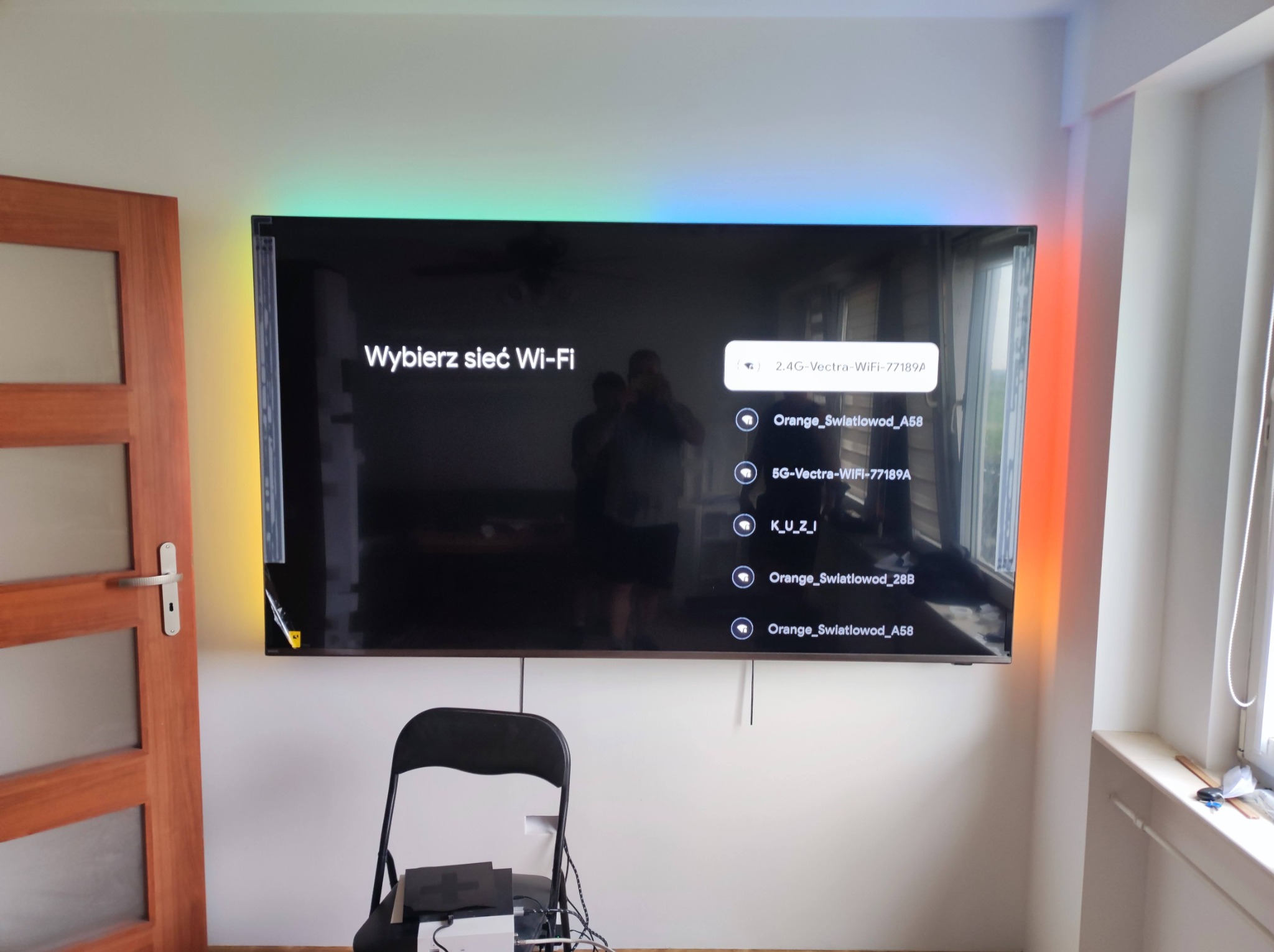 Telewizor LED z podświetleniem RGB zamontowany na ścianie, widoczny ekran z listą dostępnych sieci Wi-Fi, poniżej krzesło i sprzęt elektroniczny.