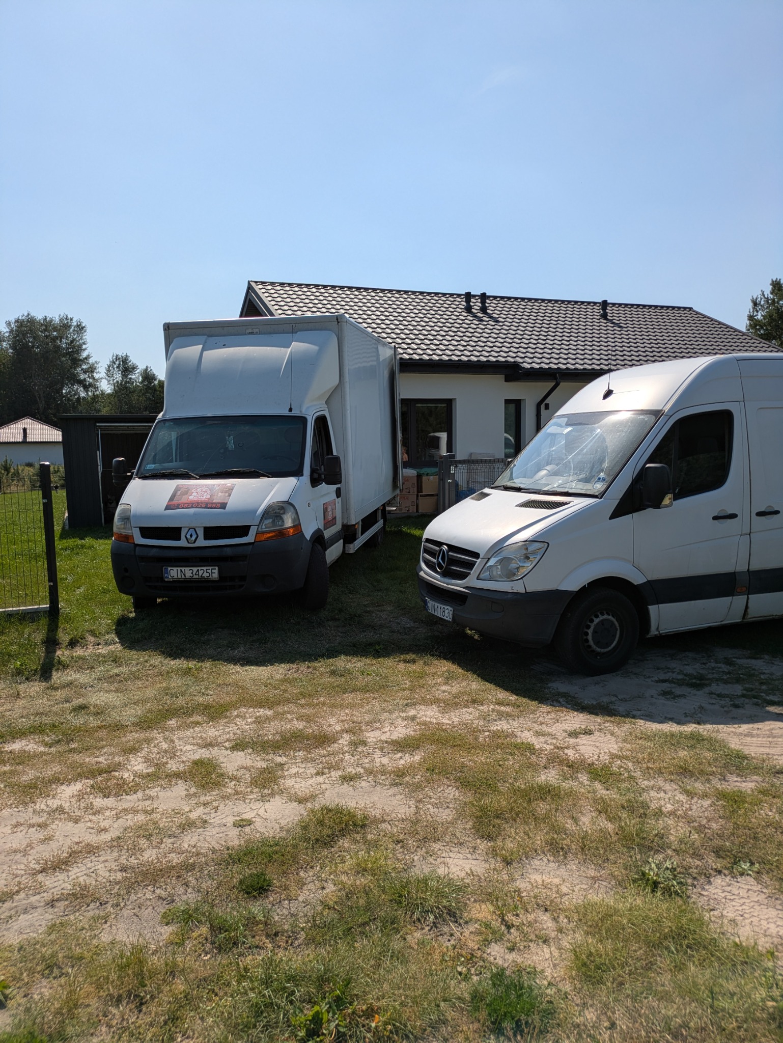 Dwa białe busy transportowe zaparkowane na posesji przed domem jednorodzinnym w słoneczny dzień. Jeden bus Renault Master, drugi Mercedes Sprinter.