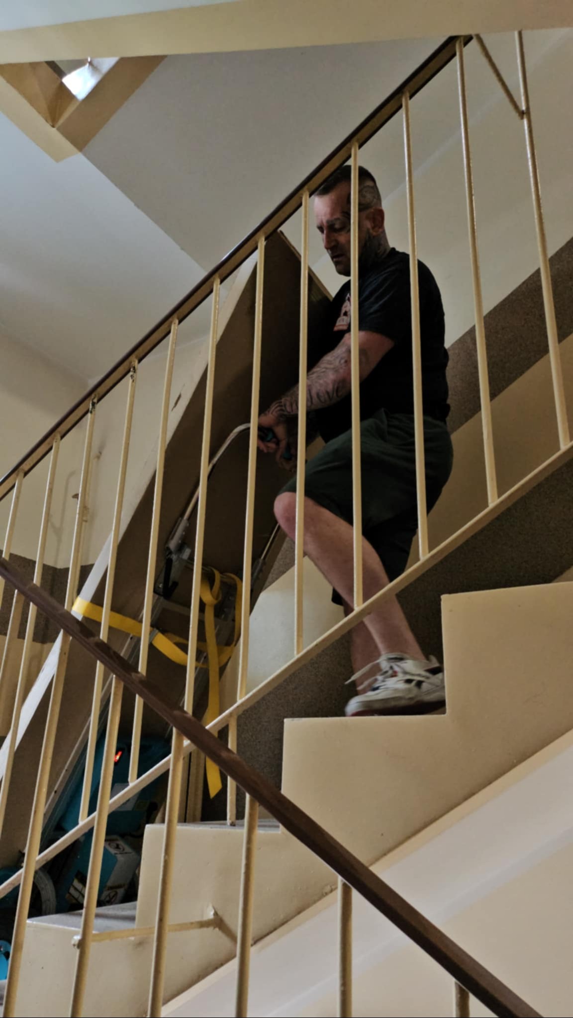 Mężczyzna z tatuażami wnosi kartonowe pudło po schodach, używając wózka schodowego z żółtymi pasami. Widok z dołu, barierki schodowe w kadrze.