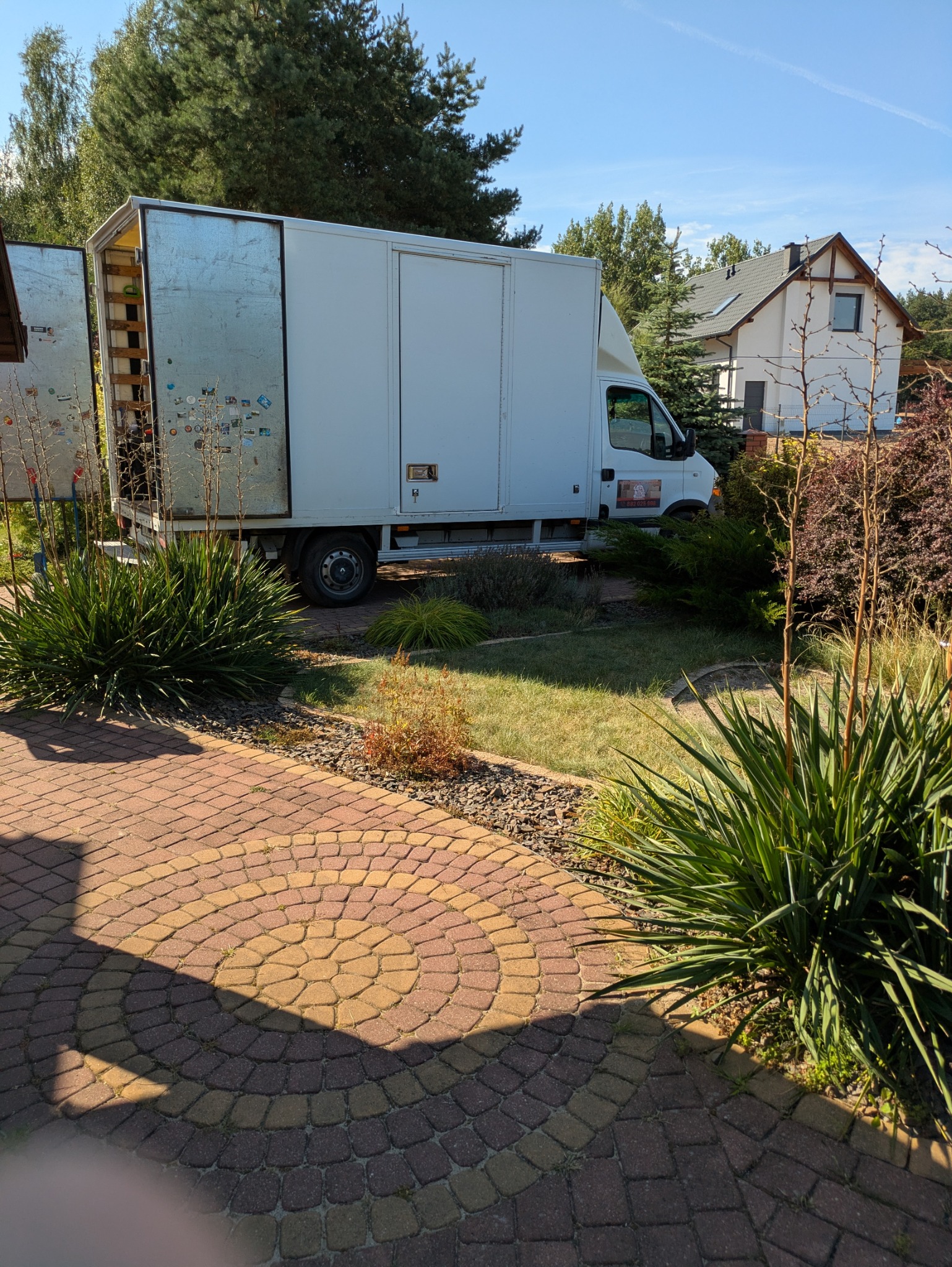 Biały samochód dostawczy z otwartą przestrzenią ładunkową, zaparkowany na trawniku przed domem jednorodzinnym w słoneczny dzień. Widoczny bruk i roślinność.