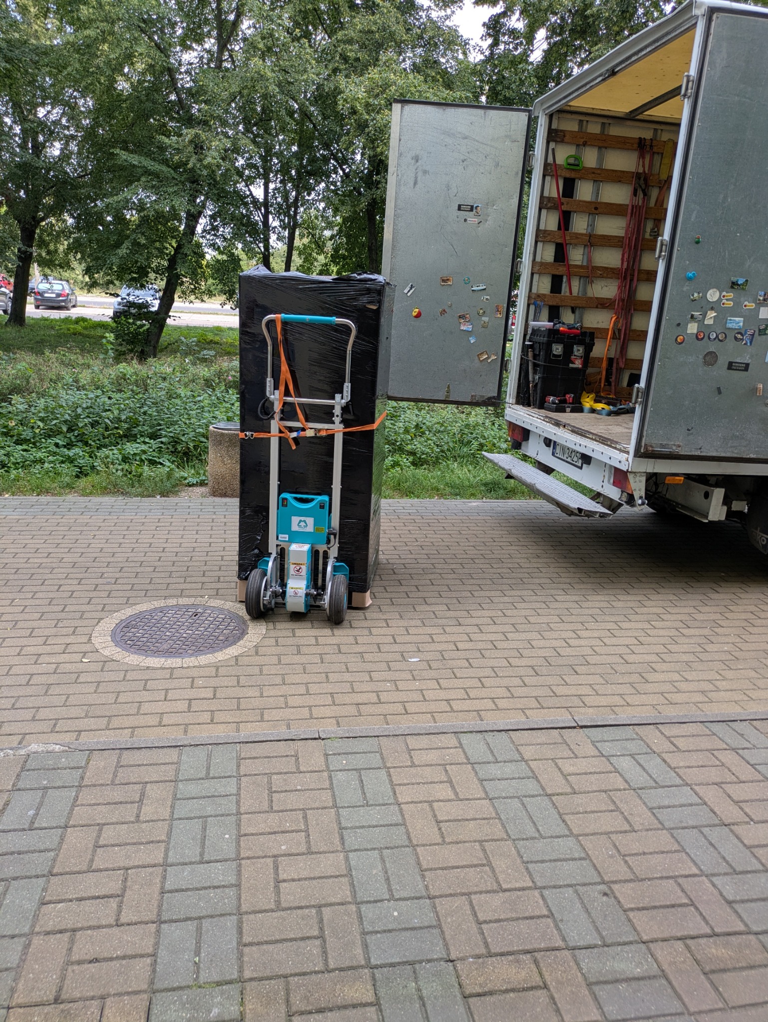 Paczka owinięta folią na wózku transportowym, obok otwartego busa na chodniku z kostki. Widoczne drzewa w tle. Detal: naklejki na drzwiach busa.