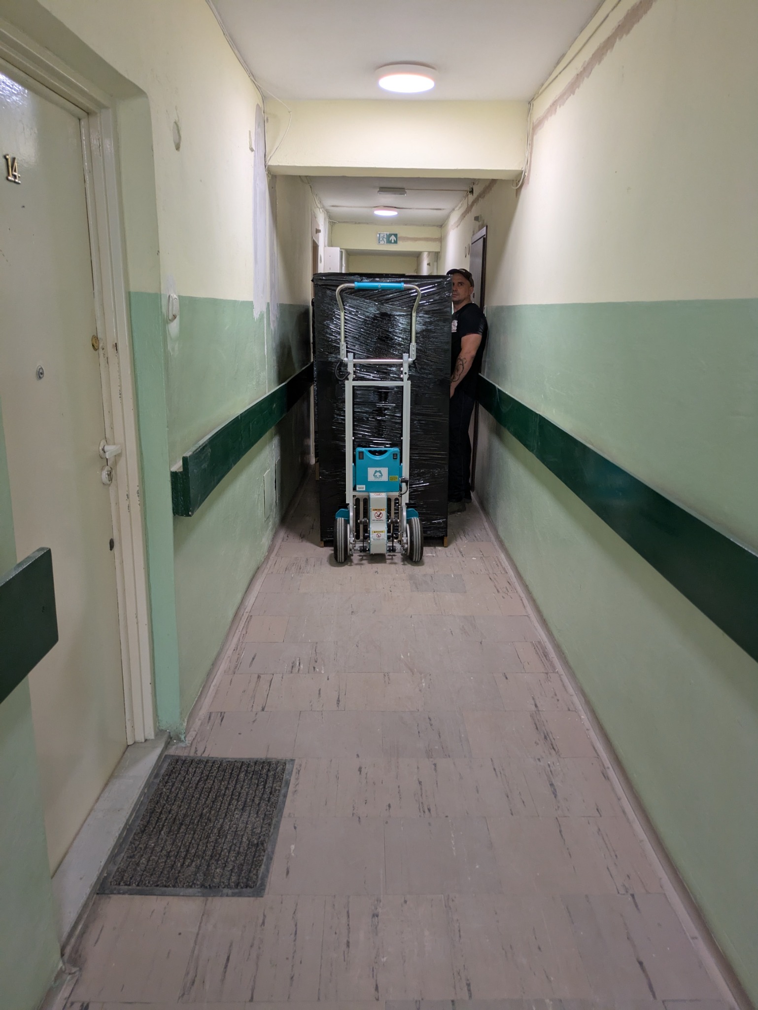 Pracownik transportuje zabezpieczony mebel wózkiem po korytarzu z numerem '14' na drzwiach. Uwagę zwraca nietypowy kolor ścian i podłogi.
