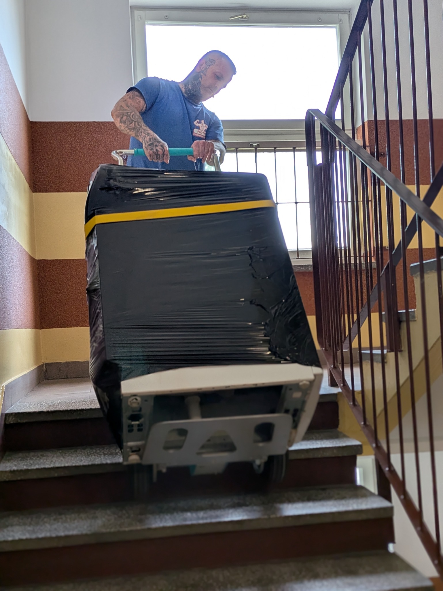 Mężczyzna z tatuażami transportuje pralkę po schodach, zabezpieczoną folią i taśmą. Widok z dołu, na klatce schodowej z oknem w tle.