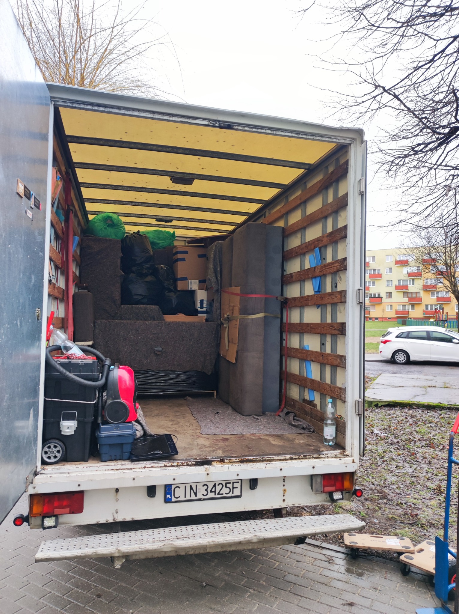 Wnętrze białego busa transportowego wypełnione meblami, kartonami i workami podczas przeprowadzki, widoczny od tyłu, z otwartymi drzwiami, tablicą rejestracyjną CIN 3425F i narzędziami do transportu.