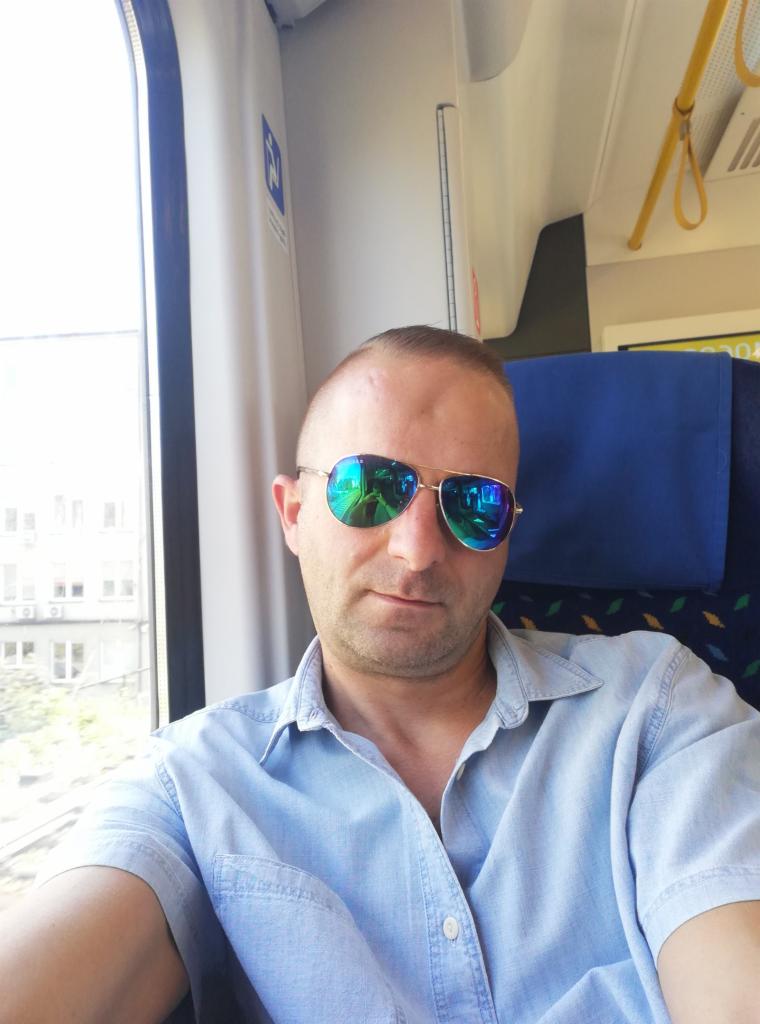 Mężczyzna w okularach z odbiciem wnętrza pociągu, siedzący przy oknie, w jasnej koszuli, na tle niebieskiego siedzenia w wagonie.