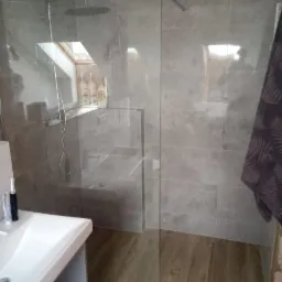 Nowoczesna kabina prysznicowa z szarymi płytkami i drewnopodobną podłogą, widoczny biały zlew i fragment okna dachowego z kosmetykami, lustrzane odbicie fotografa na szkle.