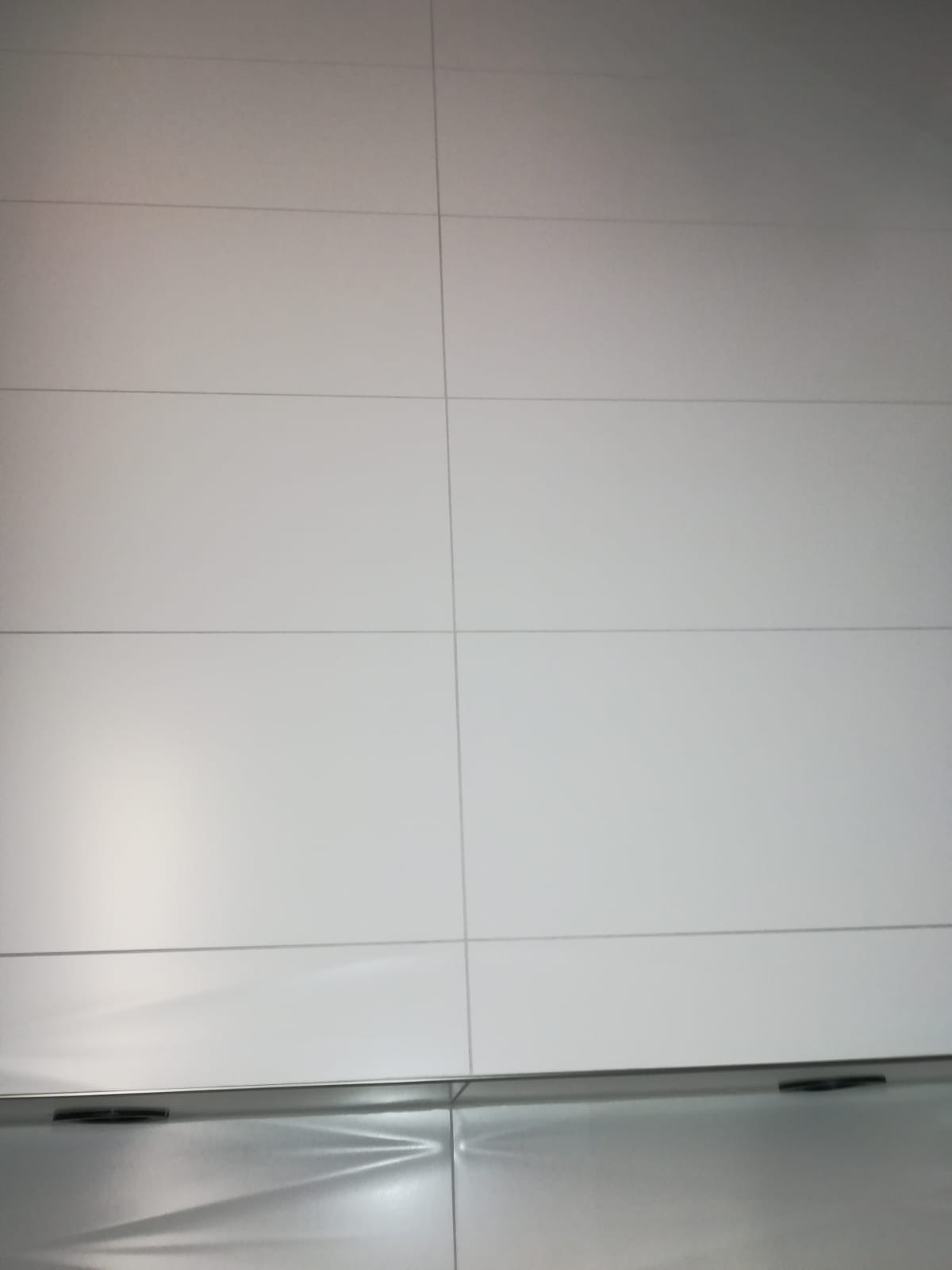 Białe, prostokątne płytki ceramiczne ułożone w rzędach z widocznymi fugami, dolna część zdjęcia ukazuje fragment metalowej powierzchni z delikatnym wzorem.