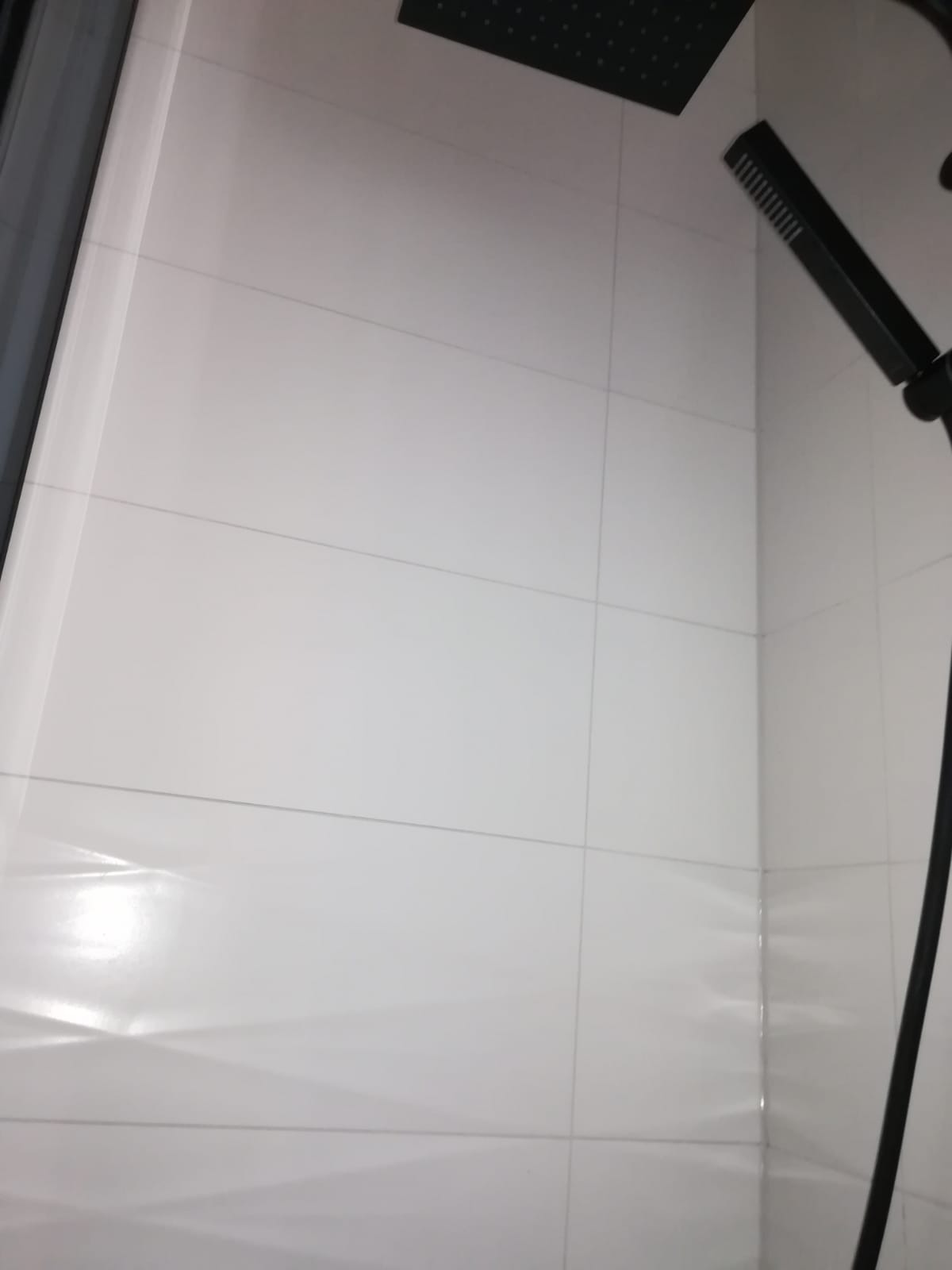 Białe, prostokątne płytki w kabinie prysznicowej, z czarną deszczownicą i baterią prysznicową w tle.