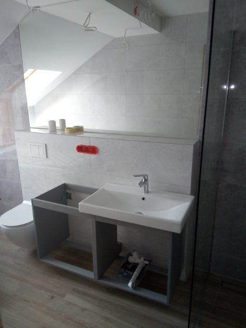 Montaż umywalki w łazience z szarymi płytkami, widoczne narzędzia i niedokończona szafka pod umywalką.