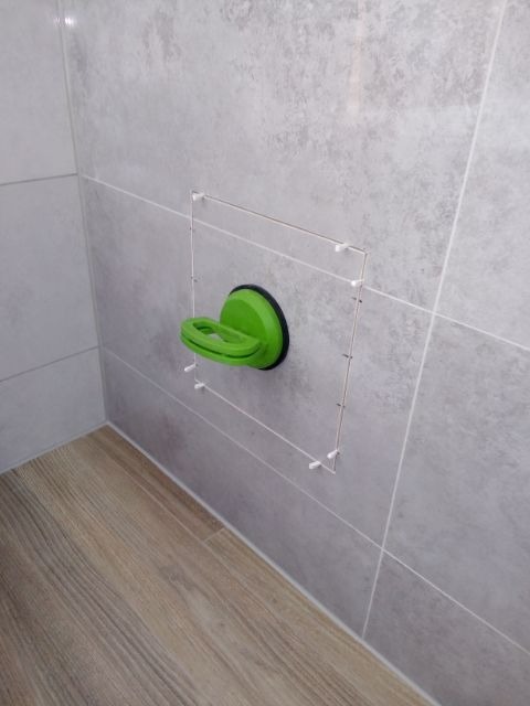 Świeżo przyklejona szara płytka ścienna, podtrzymywana przez zieloną przyssawkę, z widocznymi krzyżykami dystansowymi, na tle drewnopodobnej podłogi.