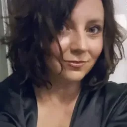 Portret uśmiechniętej kobiety z krótkimi, ciemnymi włosami i delikatnym makijażem, ubrana w ciemną, satynową bluzkę.