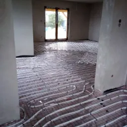 Białe rury ogrzewania podłogowego ułożone na czerwonej siatce w surowym wnętrzu z oknem, widoczne elementy mocujące.