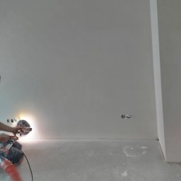 M2 Tomasz krupa - Szlifierka do gładzi w użyciu podczas prac wykończeniowych w Warszawie, widoczne gniazdka elektryczne w ścianie i ślady pyłu na podłodze.