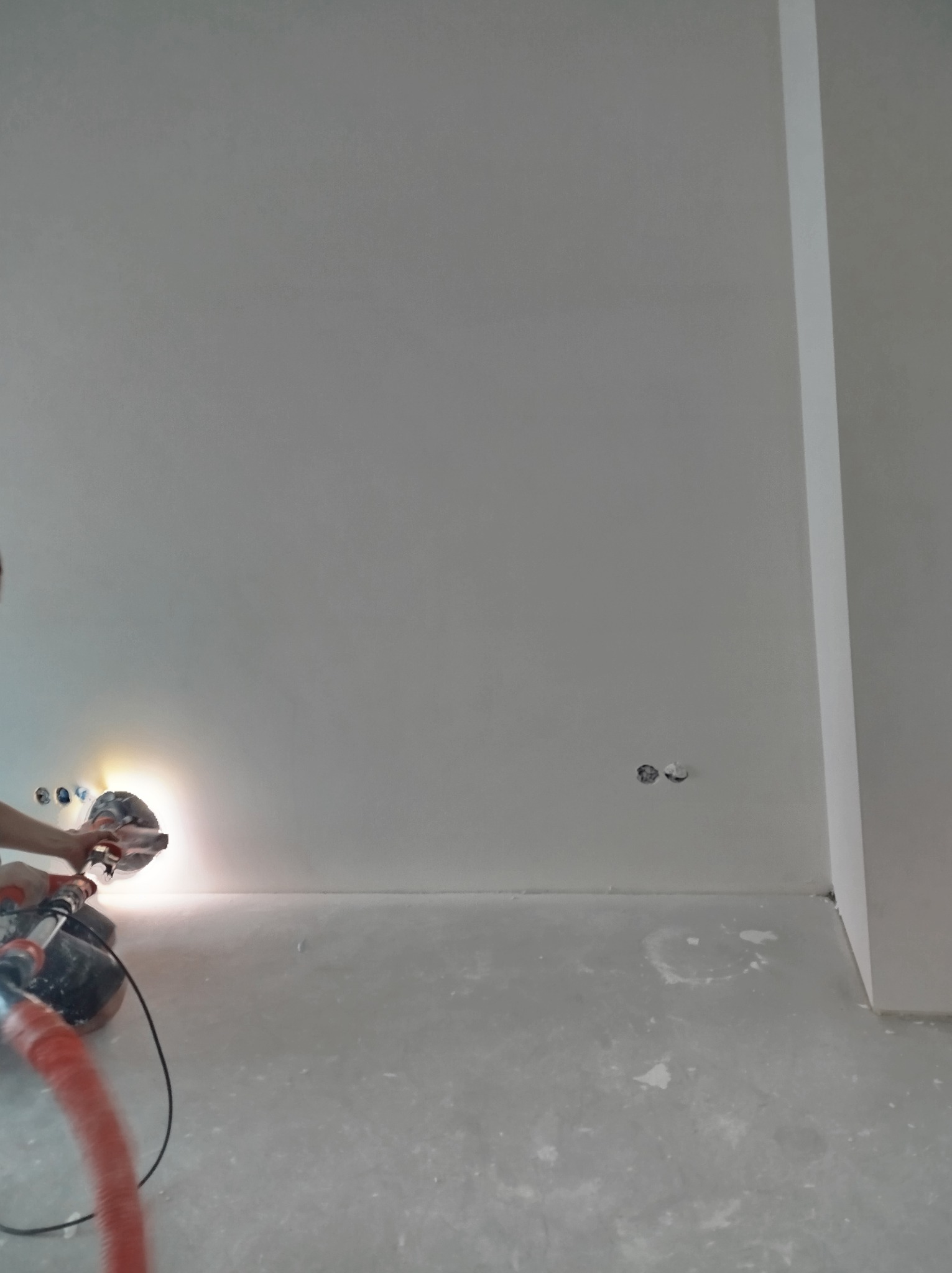 Szlifierka do gładzi w użyciu podczas prac wykończeniowych w Warszawie, widoczne gniazdka elektryczne w ścianie i ślady pyłu na podłodze.