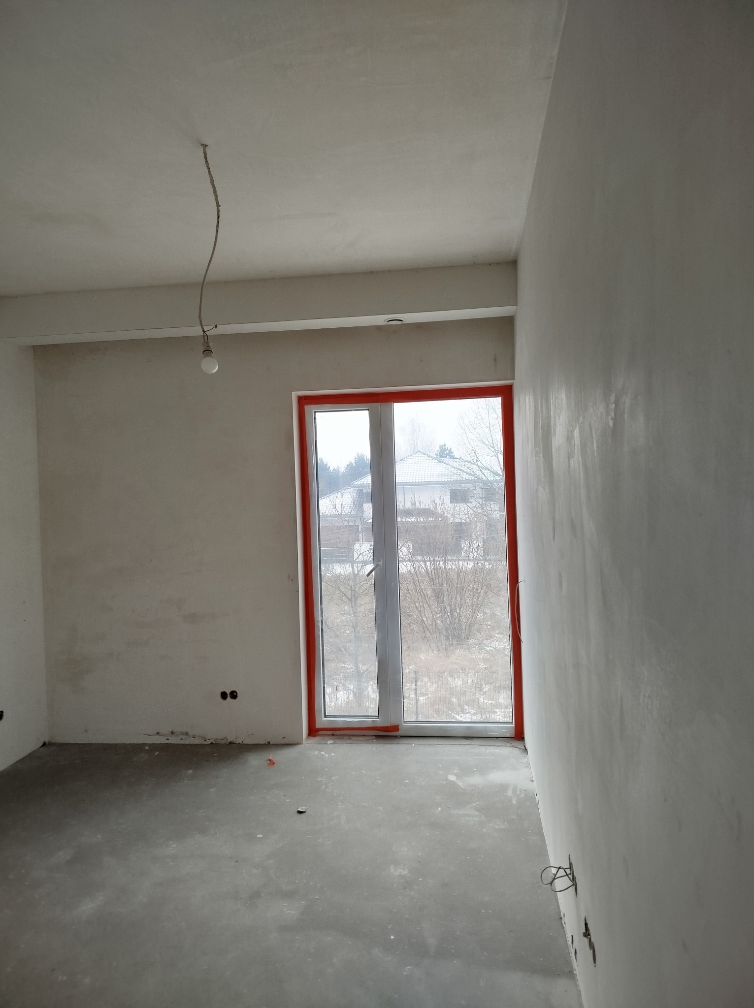 Pokój w trakcie prac wykończeniowych; ściany pokryte świeżo nałożoną warstwą, widoczne wystające przewody elektryczne, okno z pomarańczową ramą, surowy beton na podłodze.
