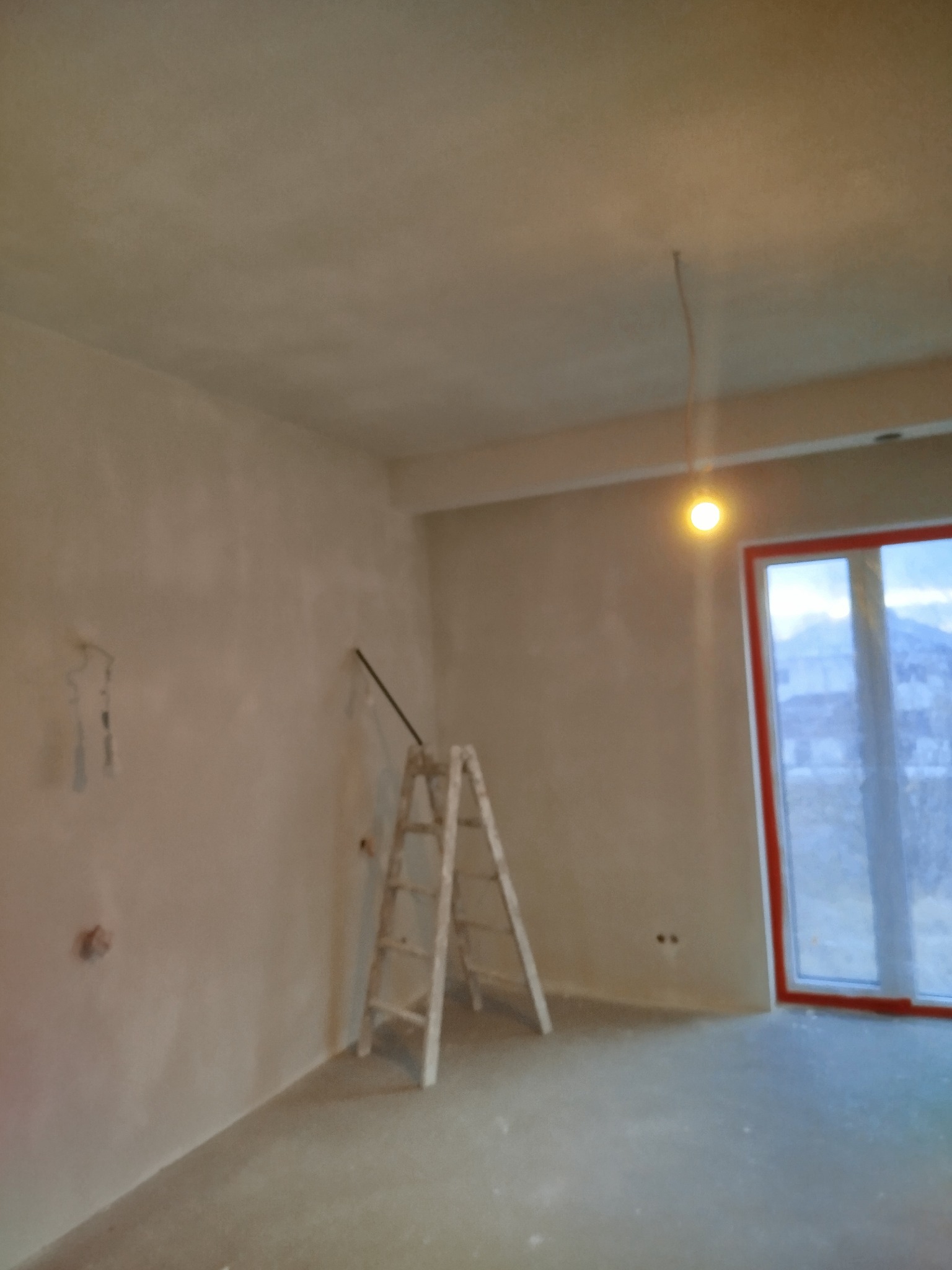 Pokój w trakcie prac wykończeniowych: ściany po nałożeniu pierwszej warstwy gładzi, widoczna drabina malarska, wystające kable elektryczne i okno z widokiem na zimowy krajobraz za szybą.