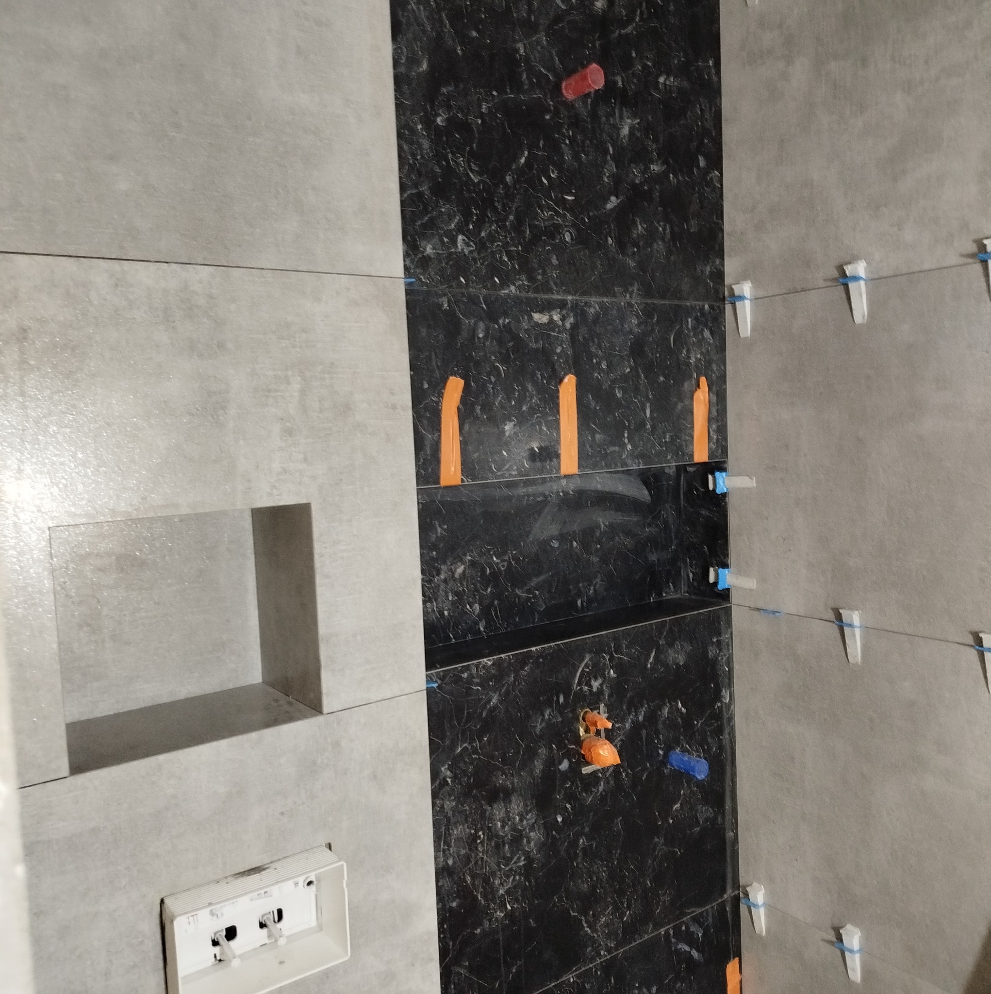 Montaż płytek w łazience: szare, matowe płytki na ścianie z wnęką i czarne płytki imitujące marmur, z użyciem poziomicy i plastikowych krzyżyków dystansowych.