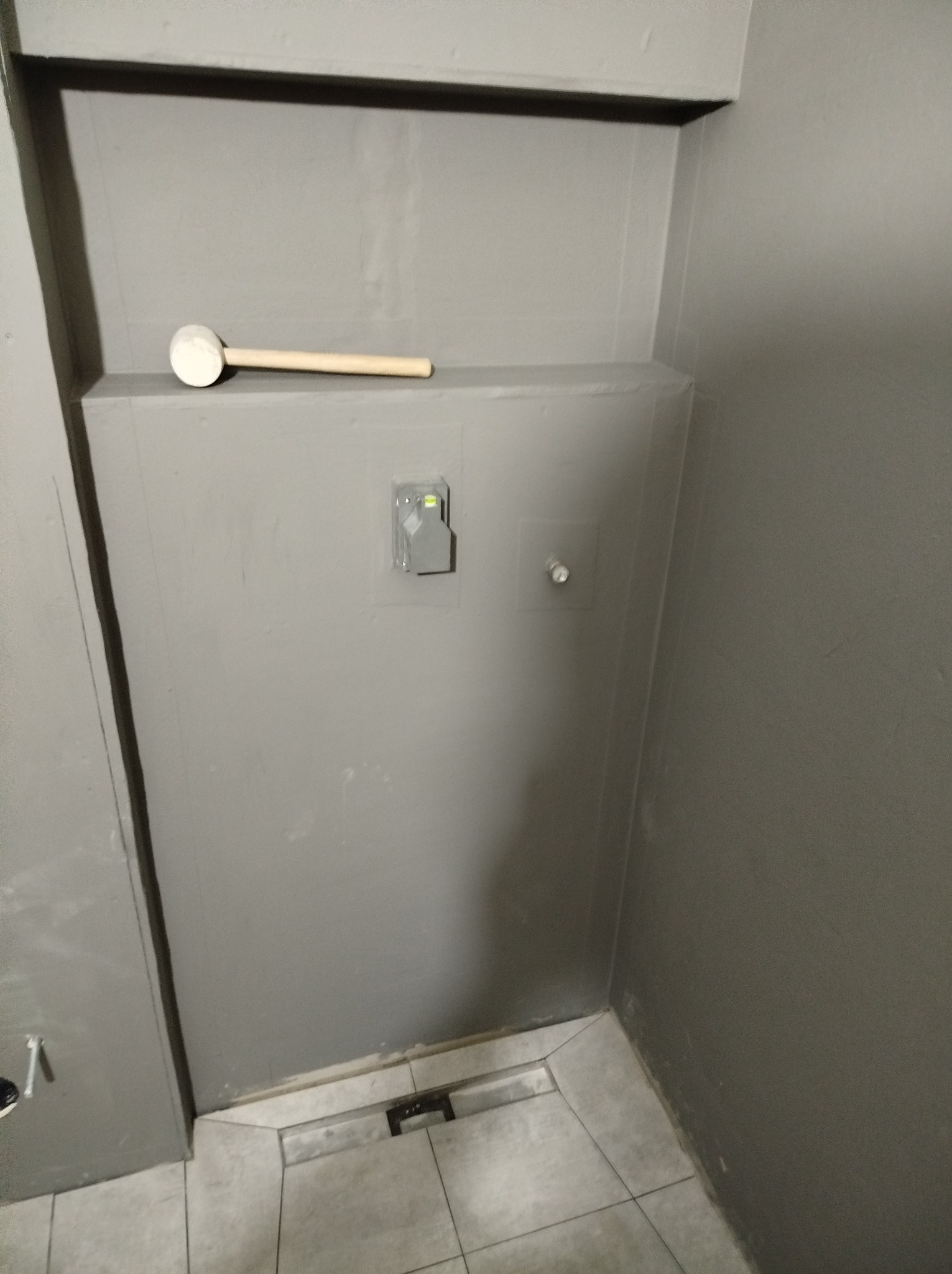 Szara ściana w trakcie prac wykończeniowych, widoczny poziomica w opakowaniu, kołek rozporowy oraz gumowy młotek na górnej krawędzi zabudowy.