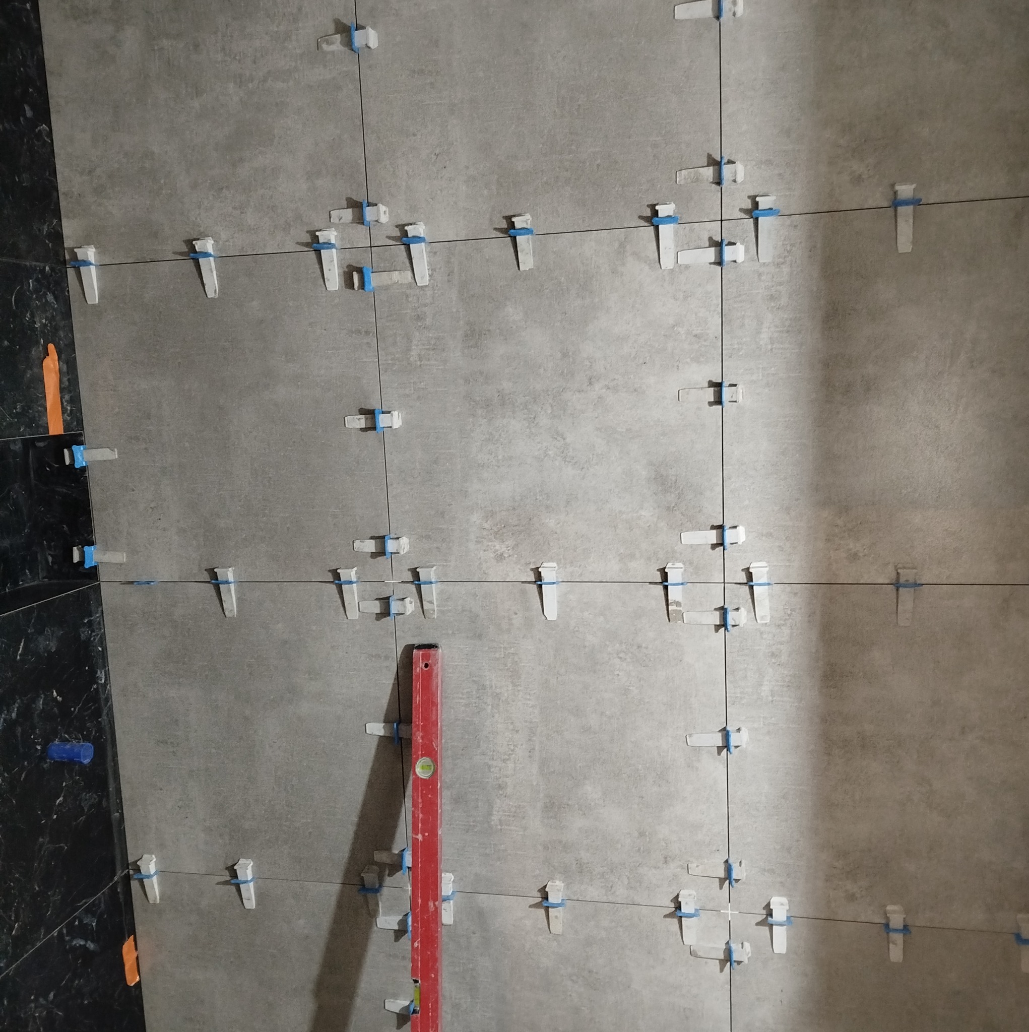 Szare płytki ścienne w trakcie układania, z użyciem systemu poziomowania płytek (klipsy i kliny z niebieskimi elementami) oraz poziomicy w kolorze czerwonym.