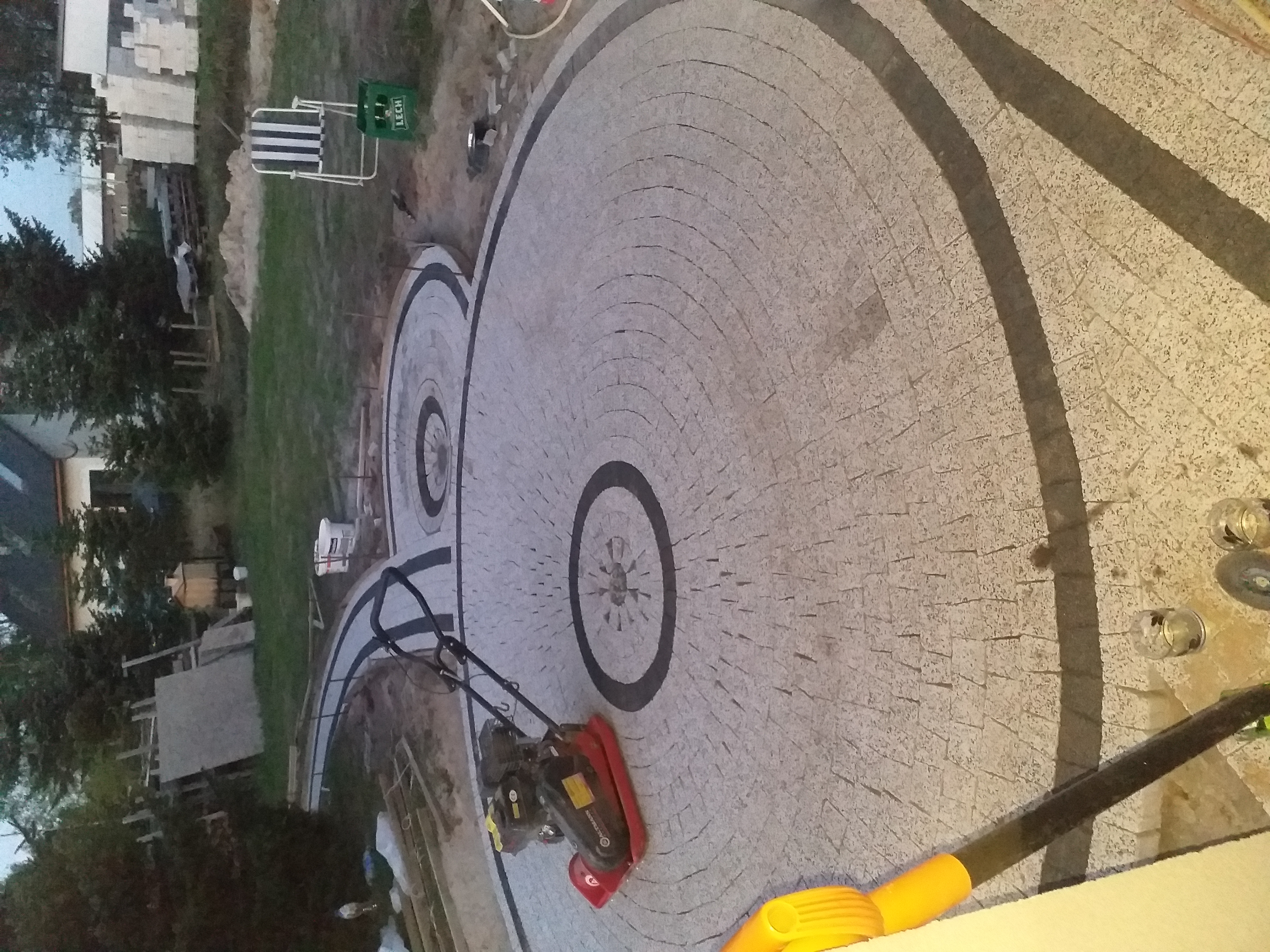 Układanie kostki granitowej w koncentryczne okręgi z dekoracyjnymi rozetami, zagęszczarka wibracyjna na powierzchni.