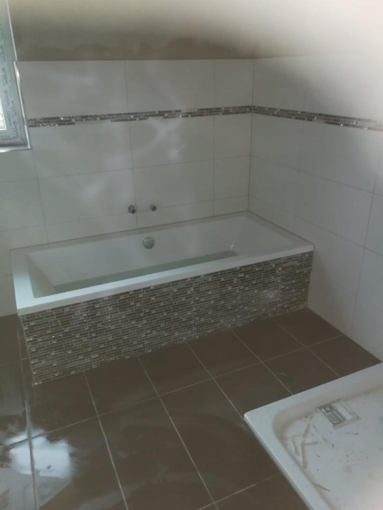 Biała wanna obłożona mozaiką w łazience z brązowymi płytkami podłogowymi i paskiem mozaiki na ścianie, widoczny fragment okna z PCV.