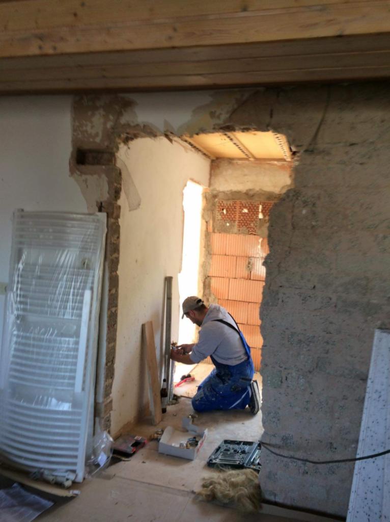 Mężczyzna w niebieskim kombinezonie klęczy w surowym wnętrzu, montując element instalacji. Widoczne cegły, nieotynkowane ściany i grzejnik w folii.