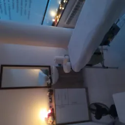 Gabinet z łóżkiem do zabiegów, widoczne białe prześcieradło, ręczniki, umywalka z lustrem, szafka i wentylator w tle. Całość oświetlona ciepłym światłem.