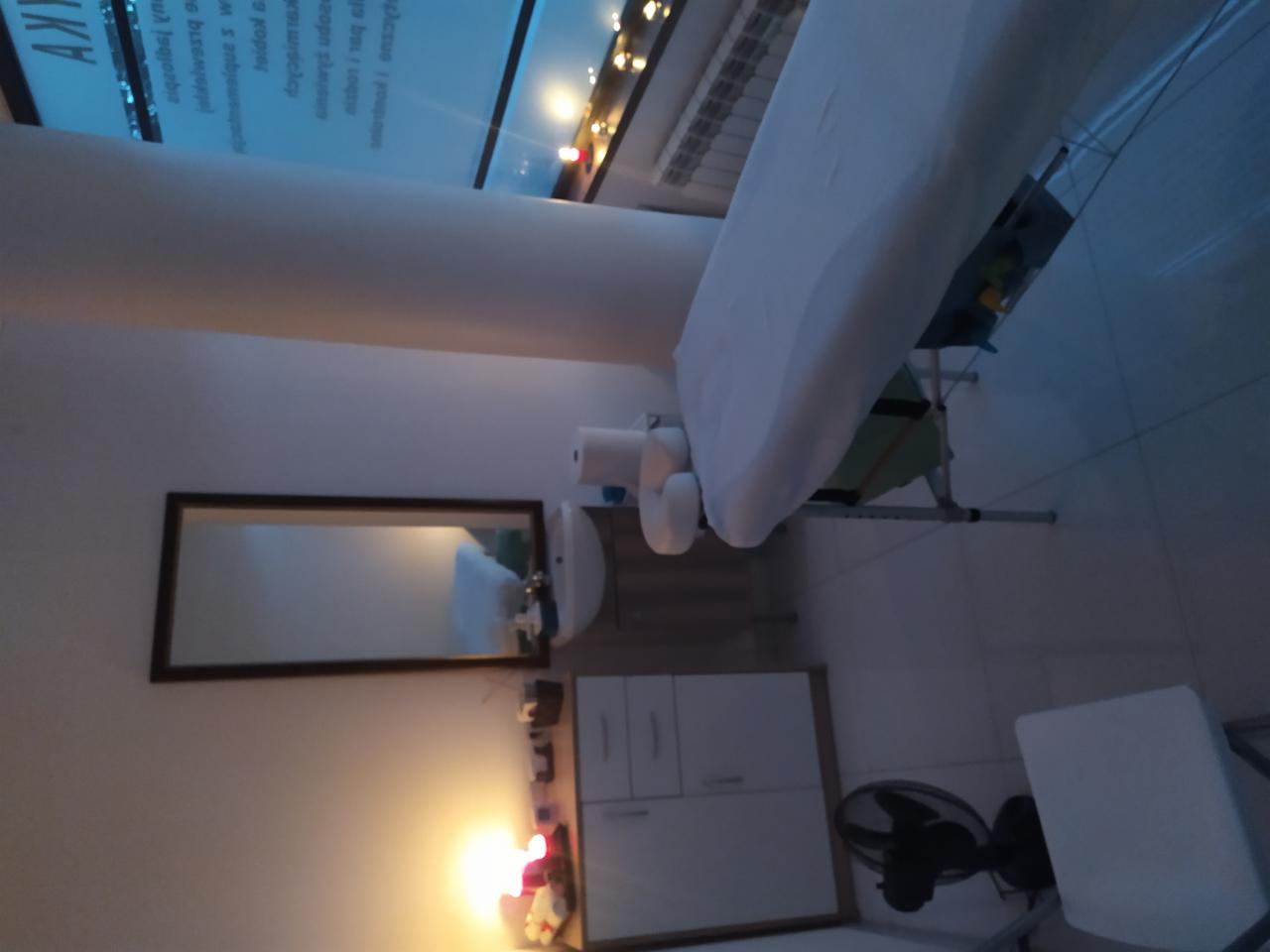 Gabinet z łóżkiem do zabiegów, widoczne białe prześcieradło, ręczniki, umywalka z lustrem, szafka i wentylator w tle. Całość oświetlona ciepłym światłem.