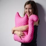 Uśmiechnięta dziewczynka tuli różową poduszkę w kształcie kota w białe kropki na tle jasnej ściany. Widoczne delikatne cienie i miękkie światło.