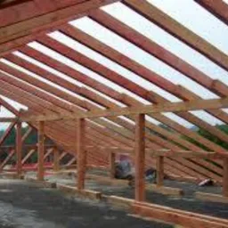 Konstrukcja dachu z belek drewnianych w surowym stanie, widok od wewnątrz podczas budowy, drabina oparta o krokwie, widoczne elementy izolacji.