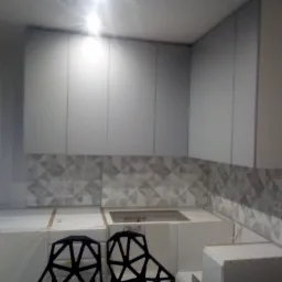 Kuchenne szafki wiszące w kolorze szarym, z geometrycznym wzorem na ścianie nad blatem, zabezpieczonym folią ochronną. Dwa czarne krzesła barowe o ażurowej konstrukcji stoją przed blatem.