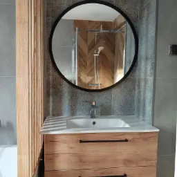 Nowoczesna łazienka z drewnianą szafką pod umywalką, okrągłym lustrem z czarną ramą odbijającym kabinę prysznicową i dekoracyjnymi płytkami ściennymi.