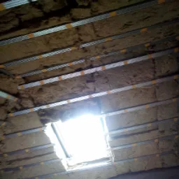 Poddasze w trakcie ocieplania wełną mineralną, widoczne profile metalowe mocowane taśmą, okno dachowe z intensywnym światłem.