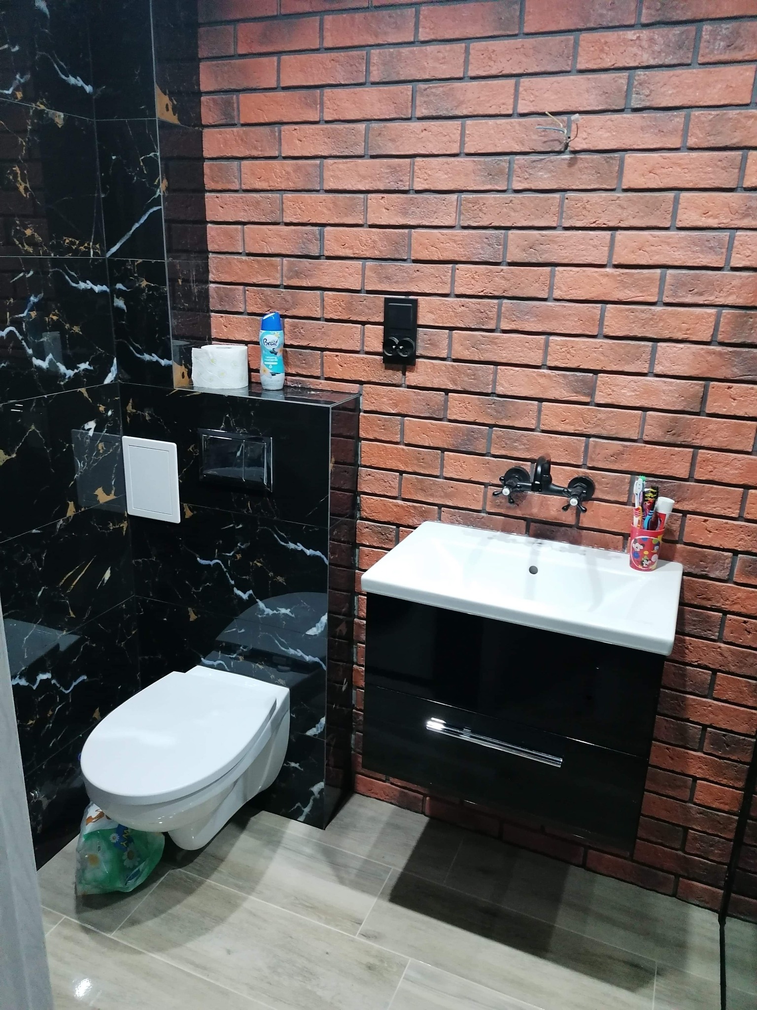 Nowoczesna łazienka z toaletą podwieszaną obudowaną czarnym marmurem, umywalką z szafką na tle ściany z cegły, widoczne wystające kable ze ściany.