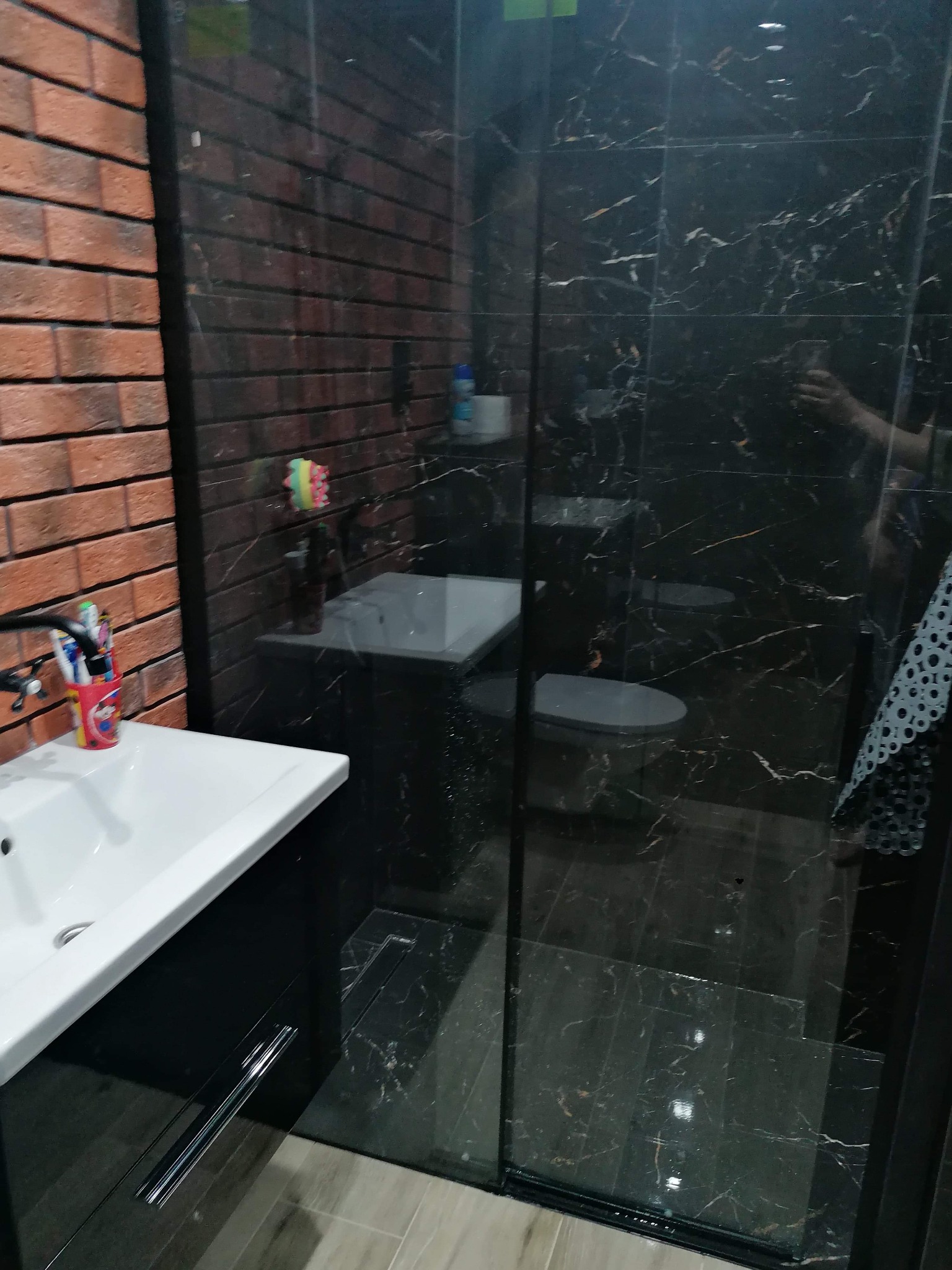 Nowoczesna łazienka z kabiną prysznicową z czarnego marmuru, umywalką na szafce i ścianą z czerwonej cegły. Widoczny odpływ liniowy pod prysznicem i akcesoria łazienkowe na umywalce.