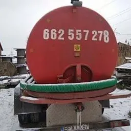 Czerwona beczka asenizacyjna z numerem telefonu na tle zimowego krajobrazu, widoczna zielona rura i drewniane szpule w tle. Zimowa sceneria.