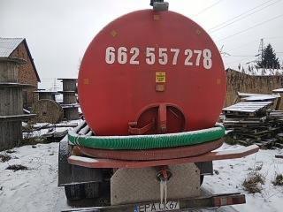 Czerwona beczka asenizacyjna z numerem telefonu na tle zimowego krajobrazu, widoczna zielona rura i drewniane szpule w tle. Zimowa sceneria.