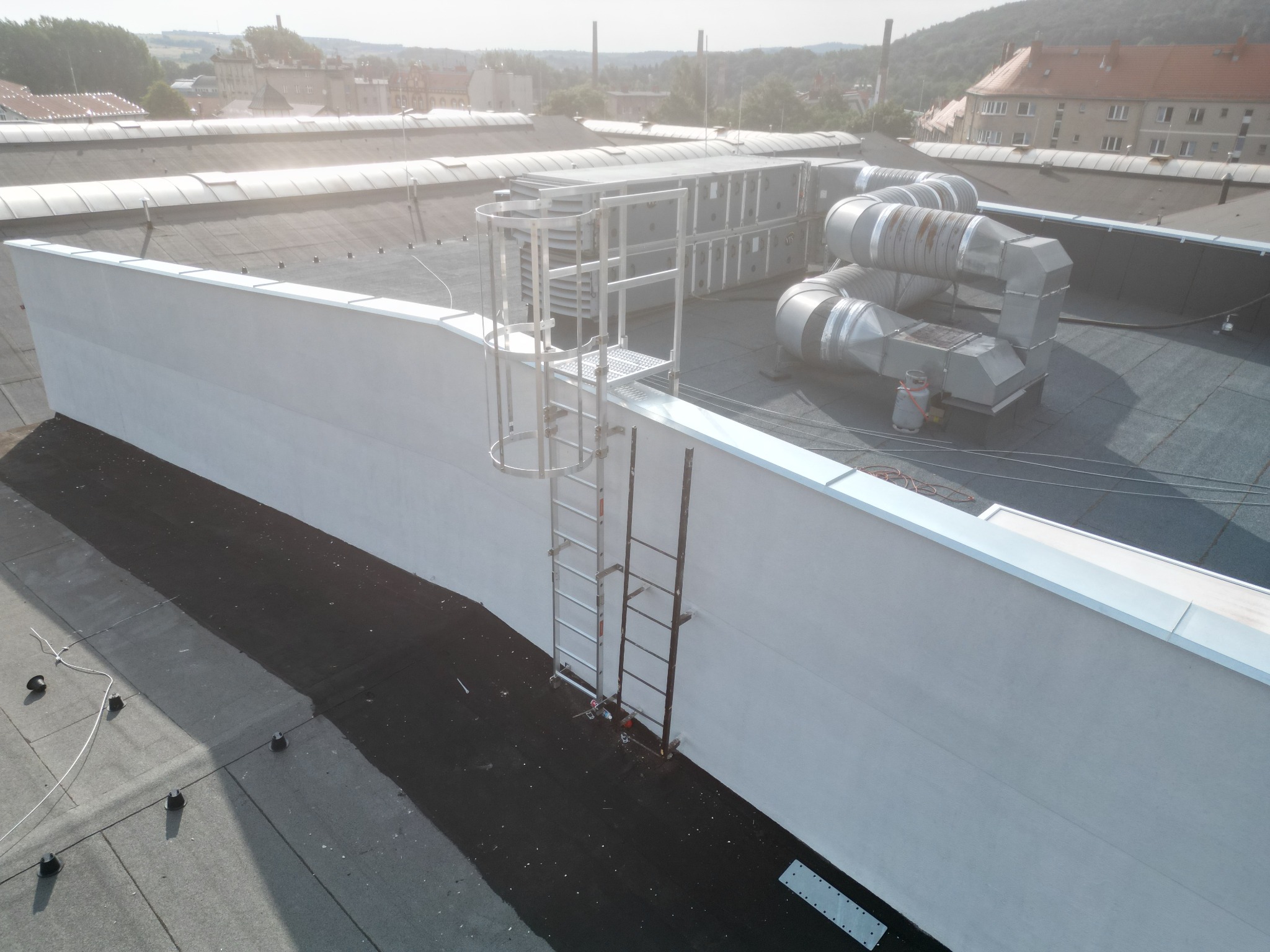Biała ściana attykowa z metalową drabinką na dachu pokrytym czarną papą. W tle widoczne instalacje wentylacyjne i budynki miejskie w lekkiej mgle.