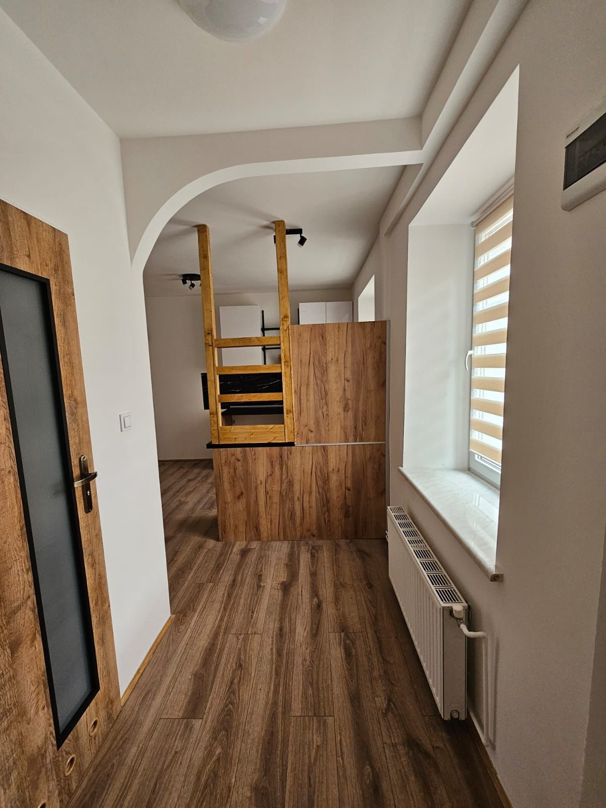 Nowoczesne wykończenie wnętrza: drewnopodobna podłoga, zabudowa kuchenna z drewnianym fornirem i czarnym blatem, widok z korytarza, grzejnik pod oknem.