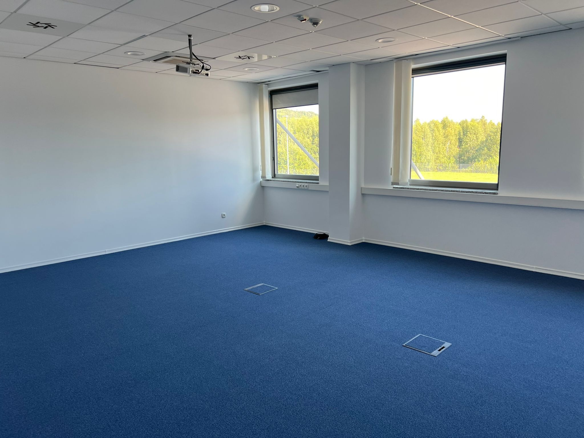Jasne biuro z niebieską wykładziną, białymi ścianami i dwoma oknami z widokiem na zieleń. Na suficie zamontowany projektor. Minimalistyczny design, czyste linie.