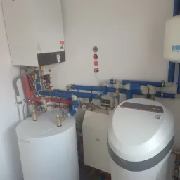 Nowoczesna kotłownia z białym piecem gazowym, zasobnikiem ciepłej wody, zbiornikiem wyrównawczym i zmiękczaczem wody, połączonymi rurami z niebieskiego i czerwonego tworzywa.