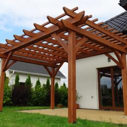 Happy Garden - Drewniana konstrukcja z belek, tworząca zadaszenie nad tarasem, z widocznym ogrodem i fragmentem domu w tle.